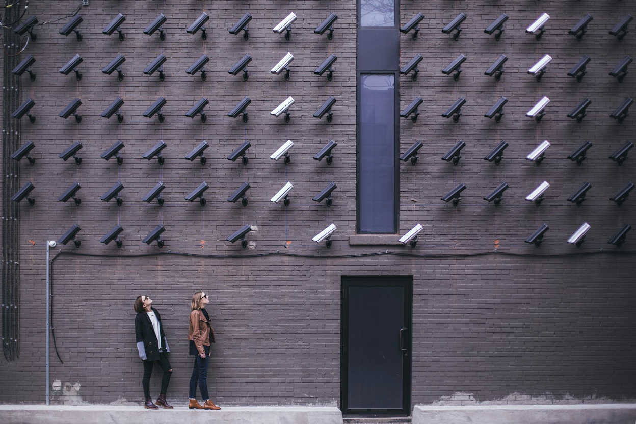 A brick wall with over 70 mounted C C T V cameras focused downward. Two people stand on the sidewalk and look up at them.
