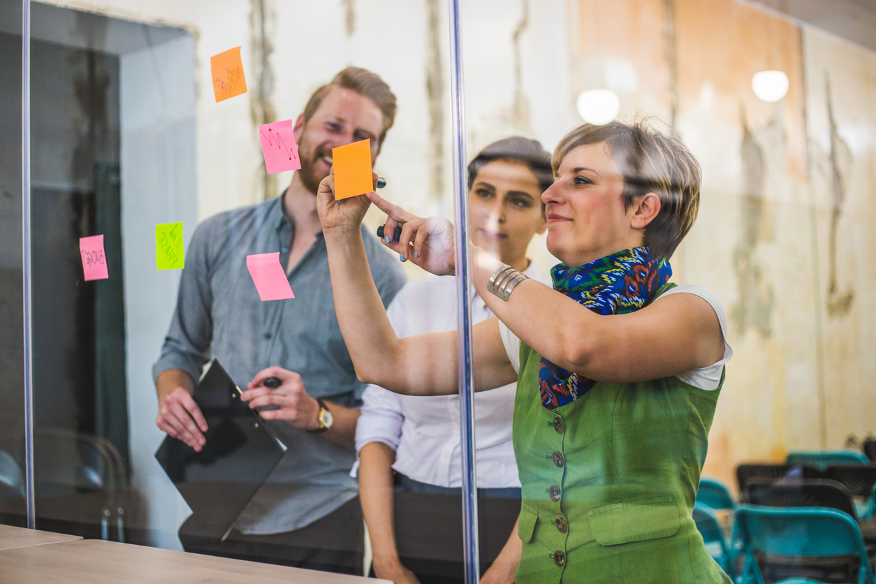Two people look on as a woman sticks a sticky note to a glass wall.