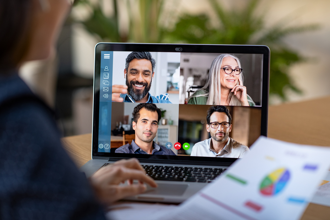 A person sits at a desk and looks at a report while four people are displayed on a video conference on a laptop on the desk.