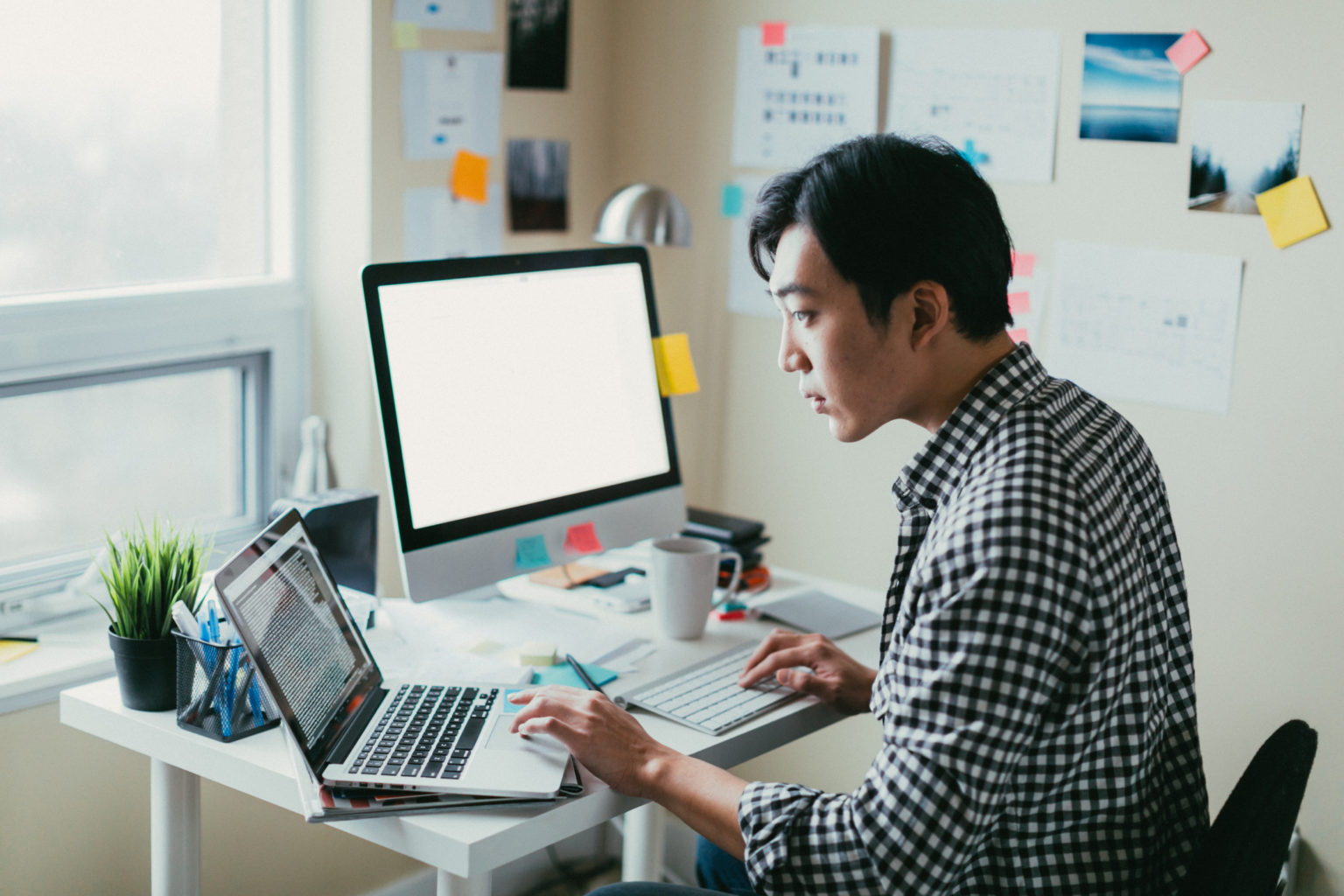 A man sits at a desk with an open laptop, a monitor, and a keyboard. He has one hand on the keyboard and the other on the mouse pad of the laptop.